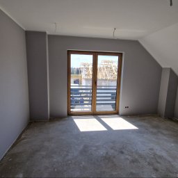 KRIS MAR - Pokój po szpachlowaniu i malowaniu na szaro, z widocznymi otworami na gniazdka elektryczne i betonową podłogą, przez okno balkonowe widać budowę.