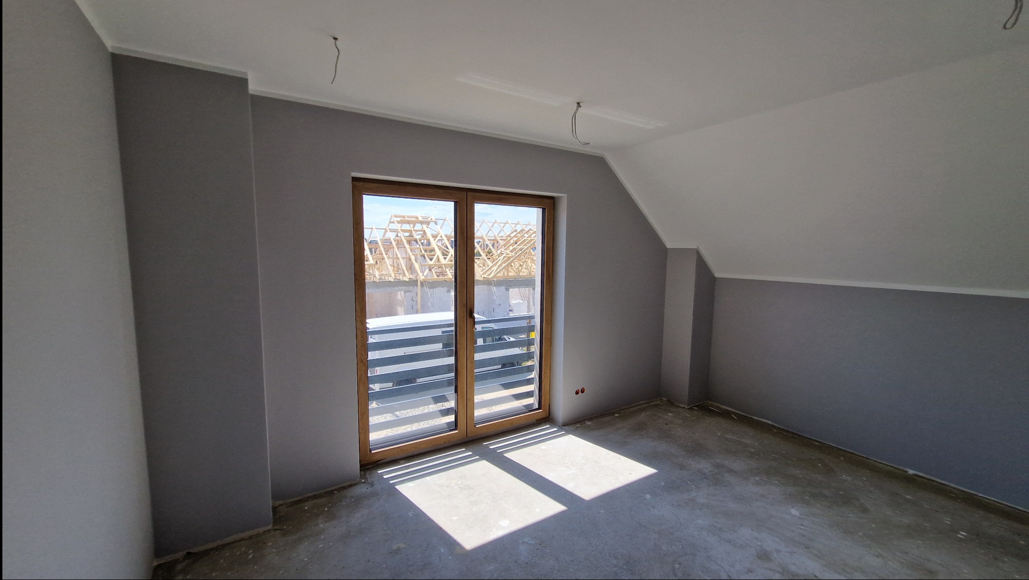 Pokój na poddaszu po malowaniu ścian na szaro, z drewnianymi drzwiami balkonowymi, surowa podłoga, widok na budowę za oknem.