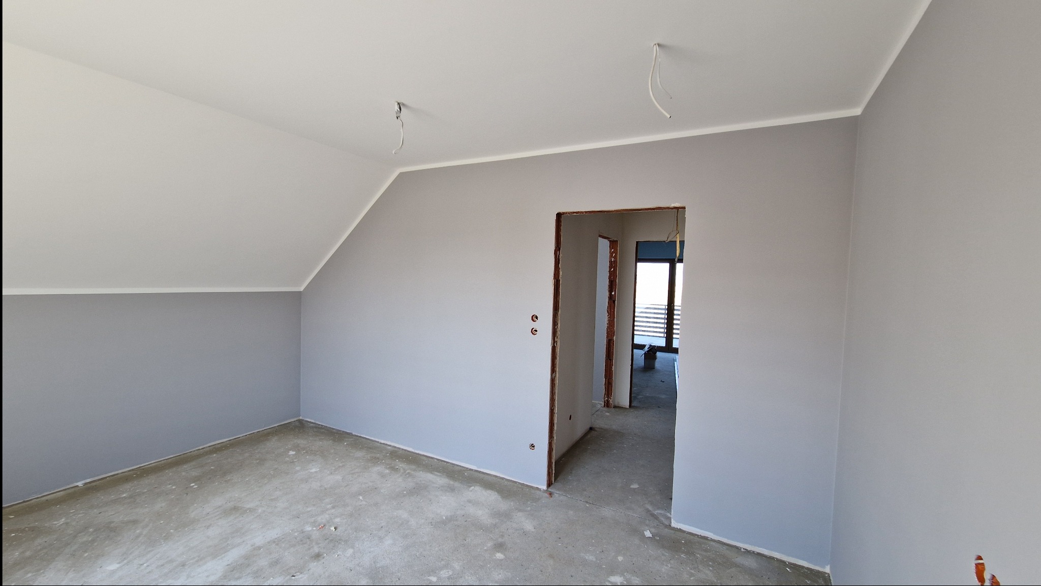 Pomalowane na szaro wnętrze poddasza ze skośnym sufitem, widoczne wystające kable elektryczne i surowa podłoga, z otwartym przejściem do kolejnego pomieszczenia.