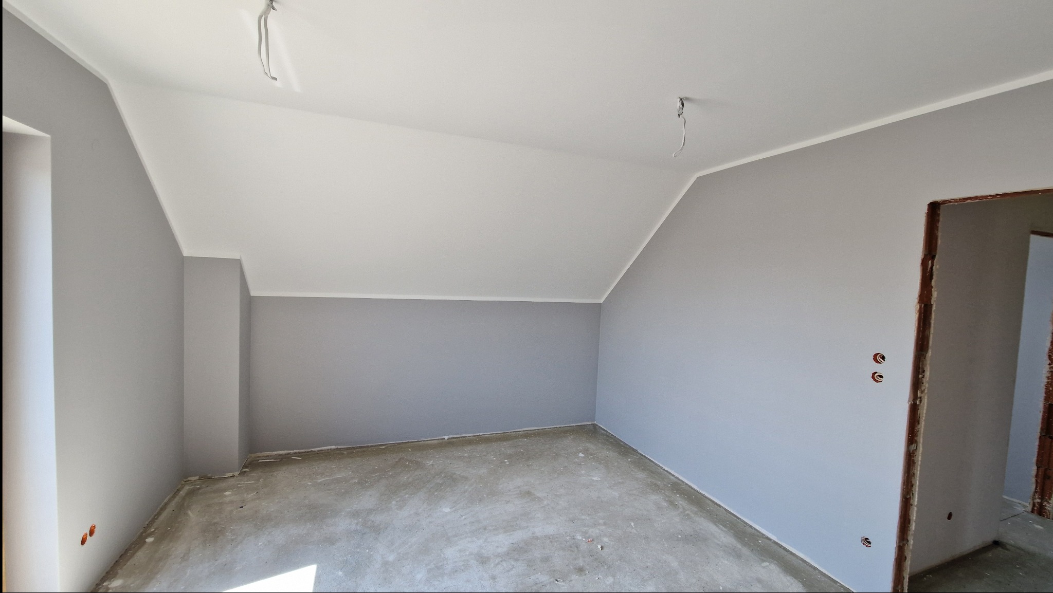 Świeżo pomalowane na szaro poddasze z białym sufitem, widoczne gniazdka elektryczne bez osprzętu i surowa podłoga przed wylewką.