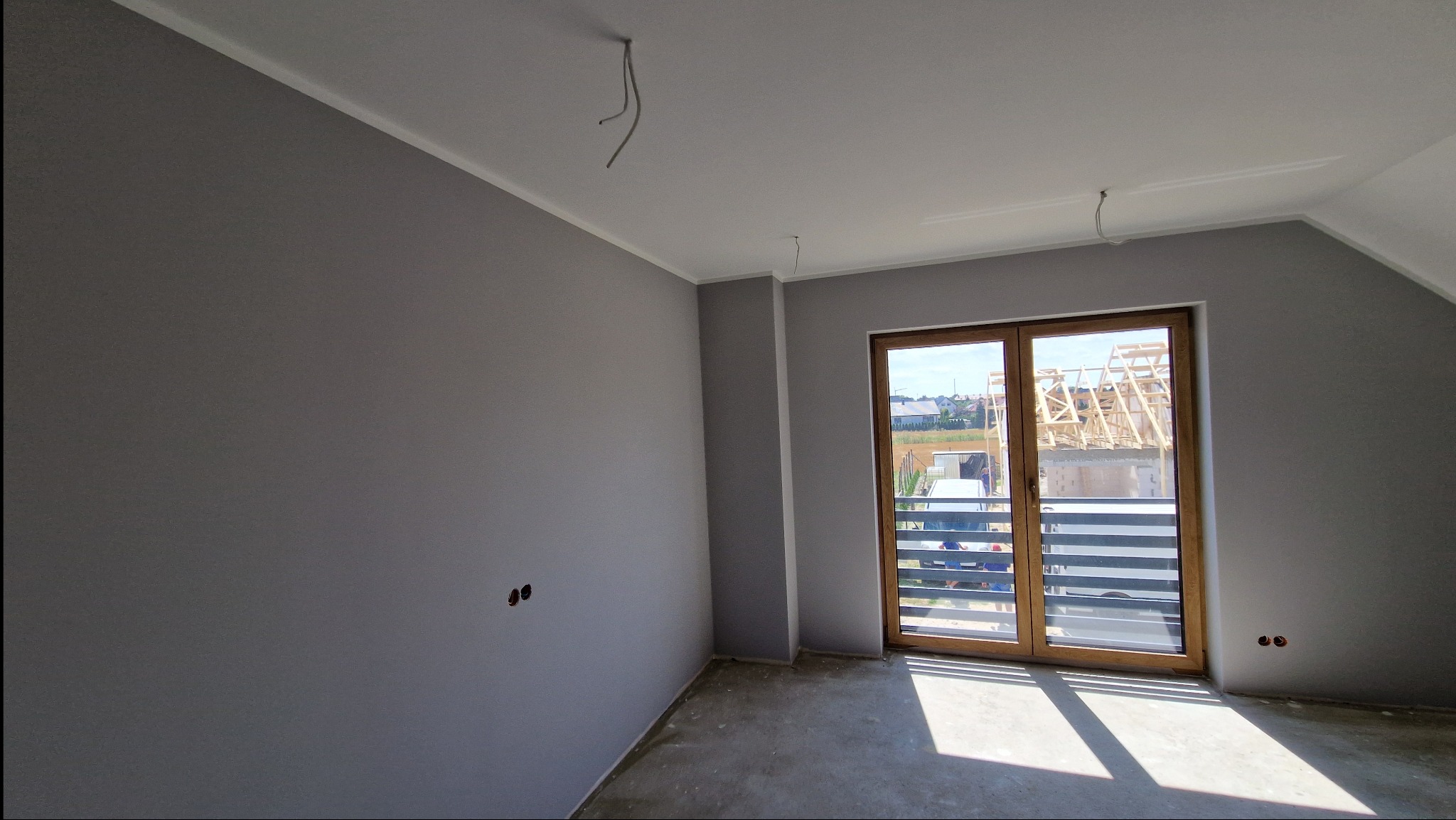 Pokój po malowaniu na szaro, widoczne wystające kable ze ściany i sufitu, surowa podłoga, okno balkonowe z widokiem na budowę domu.