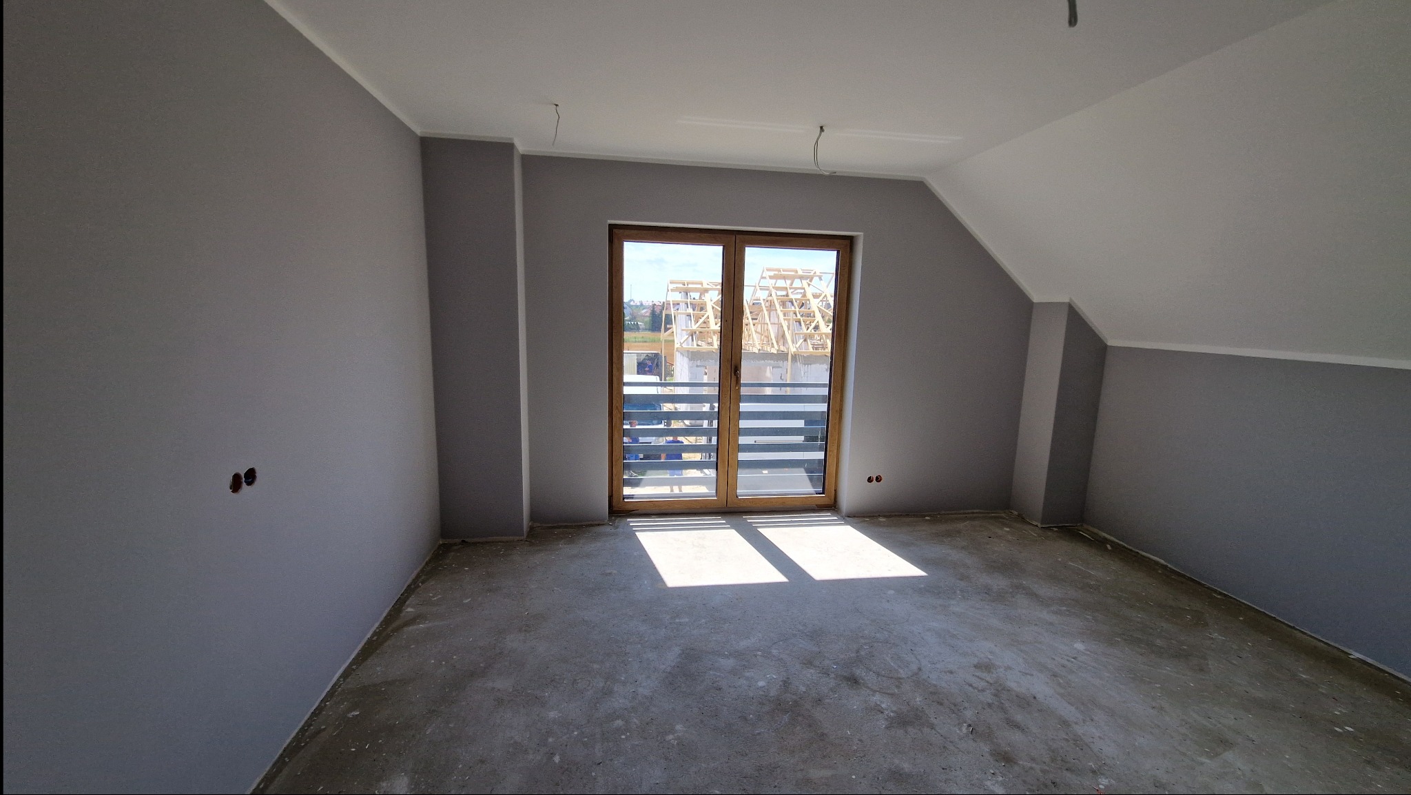 Pokój po szpachlowaniu i malowaniu na szaro, z widocznymi otworami na gniazdka elektryczne i betonową podłogą, przez okno balkonowe widać budowę.
