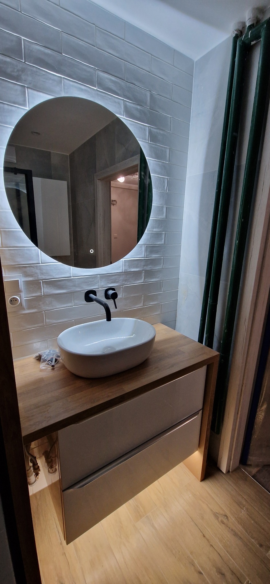 Nowoczesna łazienka z owalną umywalką nablatową i czarną armaturą na drewnianym blacie, podświetlane okrągłe lustro z wbudowanym oświetleniem LED, ściany wyłożone podłużnymi, szarymi płytkami.