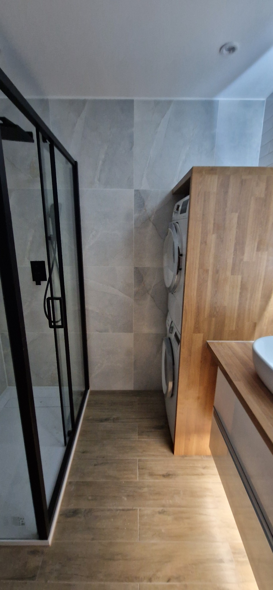 Nowoczesna łazienka z kabiną prysznicową z czarną ramą, pralką i suszarką w drewnianej obudowie oraz umywalką na drewnianym blacie.