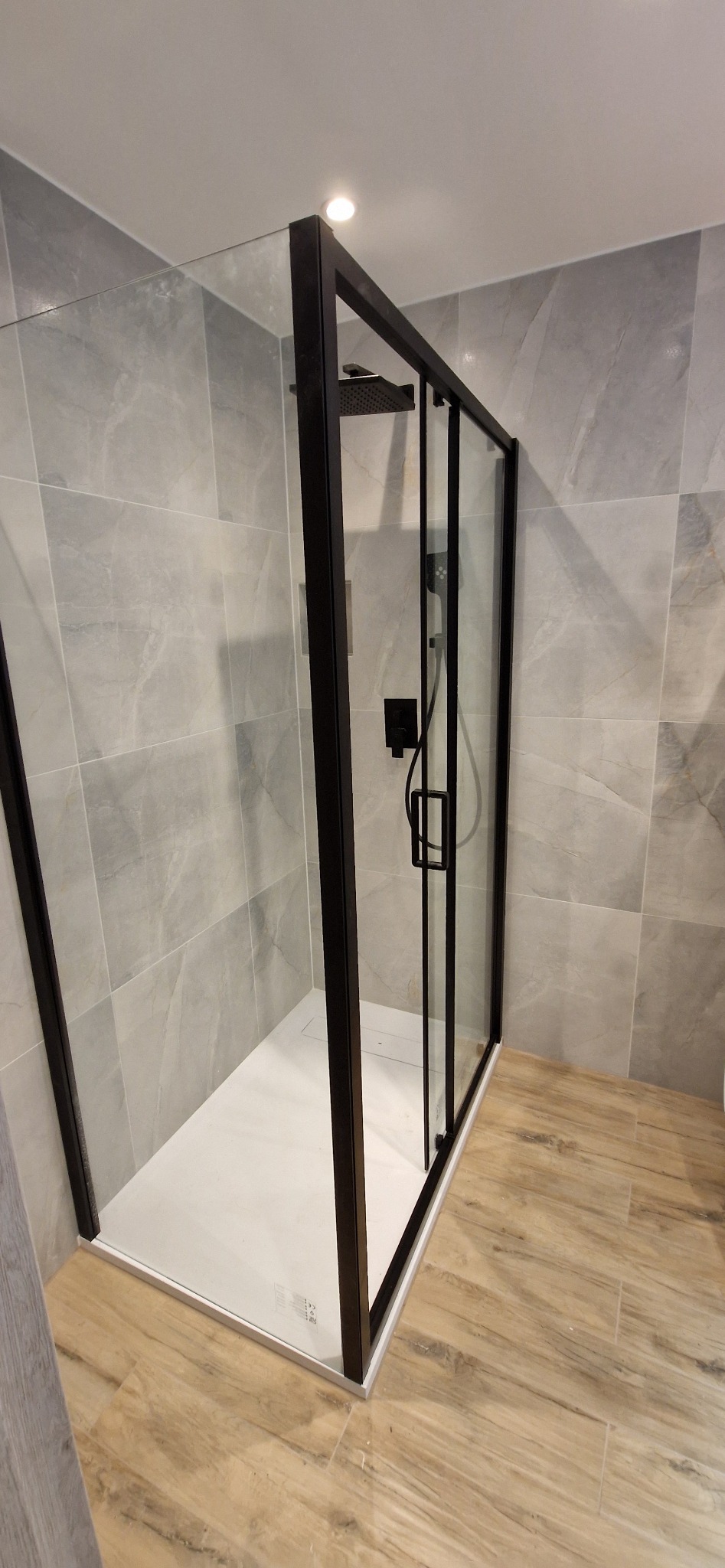 Kabina prysznicowa z czarnymi ramami i deszczownicą, wykończona szarymi płytkami imitującymi marmur na ścianach i drewnopodobnymi płytkami na podłodze.