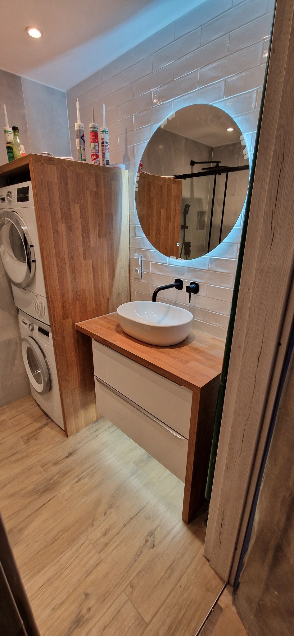 Nowoczesna łazienka z umywalką nablatową, okrągłym lustrem z podświetleniem LED, drewnianym blatem i pralką w zabudowie. Na blacie widoczne silikony sanitarne.