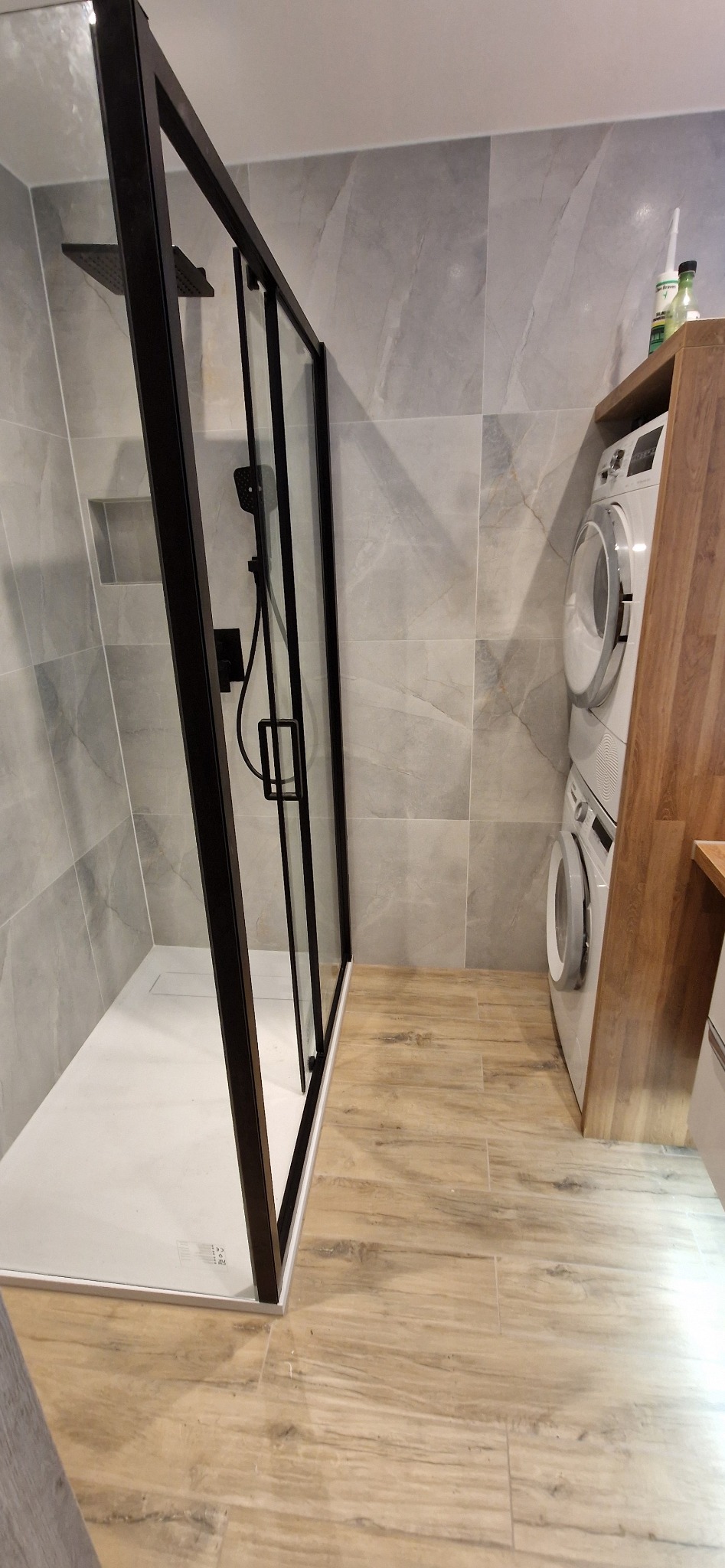 Nowoczesna łazienka z kabiną prysznicową typu walk-in z czarną ramą, szarymi płytkami imitującymi marmur na ścianach i drewnopodobnymi płytkami na podłodze, obok zabudowana pralka i suszarka.