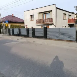 Nowoczesne ogrodzenie panelowe w kolorze szarym z czarnymi słupkami, widok z ulicy na tle domu z balkonem.