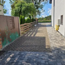 Ułożona nawierzchnia z ażurowych płyt betonowych wypełnionych kamieniem, obok chodnik z szarej kostki brukowej, drewniane ogrodzenie i brama z widocznymi śladami korozji.