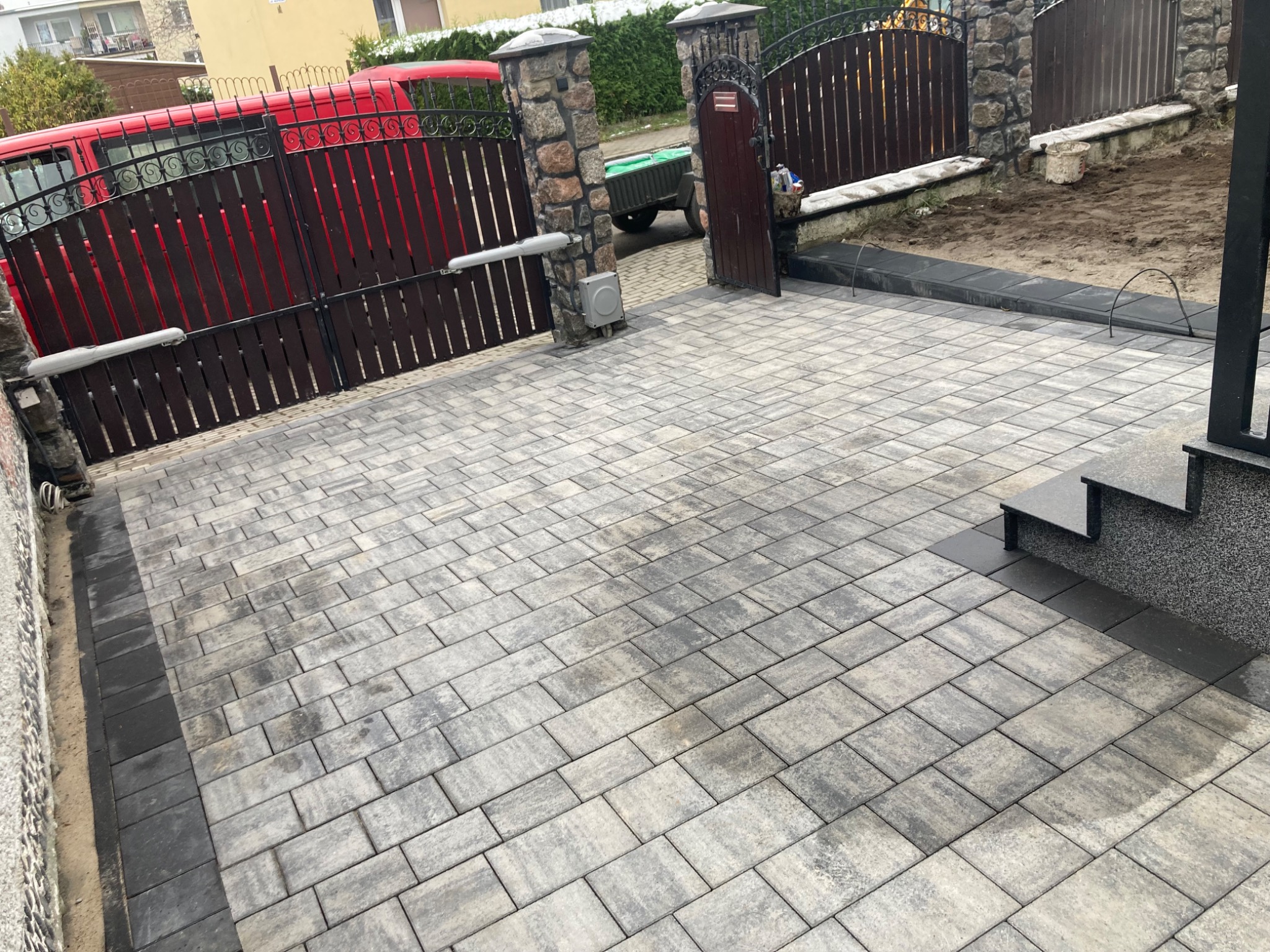 Ułożona szara kostka brukowa z ciemnoszarą obwódką, prowadząca do schodów i bramy z automatyką, z widocznym czerwonym samochodem w tle.