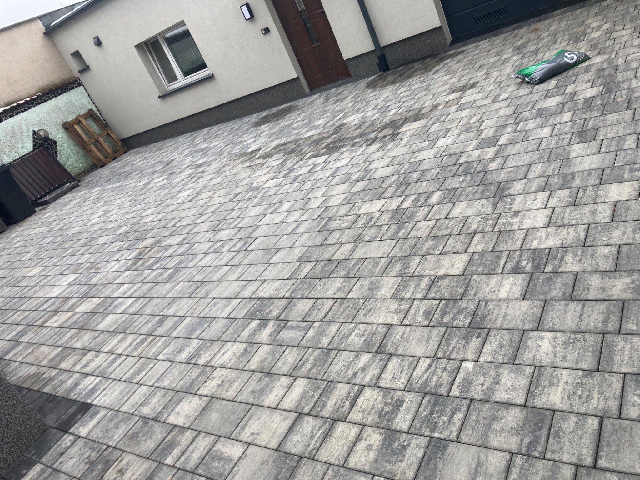 Ułożona szara kostka brukowa na podjeździe, widoczny fragment domu z oknem i drzwiami wejściowymi, oraz worek z materiałem budowlanym.