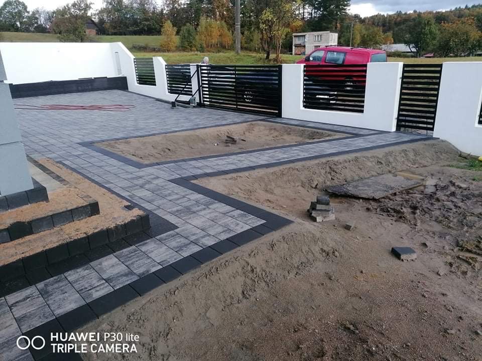 Ułożona szara kostka brukowa z ciemnymi obrzeżami na podjeździe, widoczny fragment schodów i przygotowane miejsce na trawnik, w tle białe ogrodzenie i czarna brama.