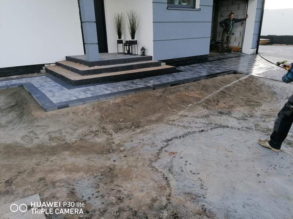 Fragment układanej kostki brukowej przed domem z widocznymi schodami i przygotowanym podłożem z piasku. W tle widoczna osoba w drzwiach.