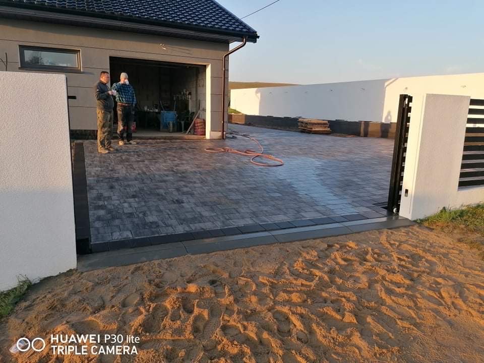 Ułożona kostka brukowa na podjeździe do garażu, widoczni dwaj mężczyźni w tle, biały mur z bramą wjazdową po prawej stronie, piasek na pierwszym planie.