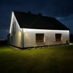 Nowoczesny dom jednorodzinny oświetlony nocą białym światłem LED, podświetlony dach i elewacja. Widok z ogrodu, ciemne niebo w tle, zielony trawnik.