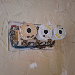 ELMIG BARTOSZ SKOWROŃSKI - Gniazdo bezpiecznikowe z trzema bezpiecznikami topikowymi na ścianie o nierównej fakturze. Widoczne przewody elektryczne i elementy mocujące.