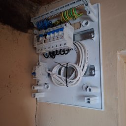 ELMIG BARTOSZ SKOWROŃSKI - Rozdzielnica elektryczna z bezpiecznikami, przewodami i listwą zaciskową zamontowana na ścianie. Widoczne kable w kolorach: niebieskim, zielono-żółtym i brązowym.