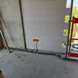 ELMIG BARTOSZ SKOWROŃSKI - Surowe ściany z instalacją elektryczną w peszlach, widoczne pomarańczowe puszki. W tle rusztowanie i budynek. Wnętrze w trakcie prac wykończeniowych.