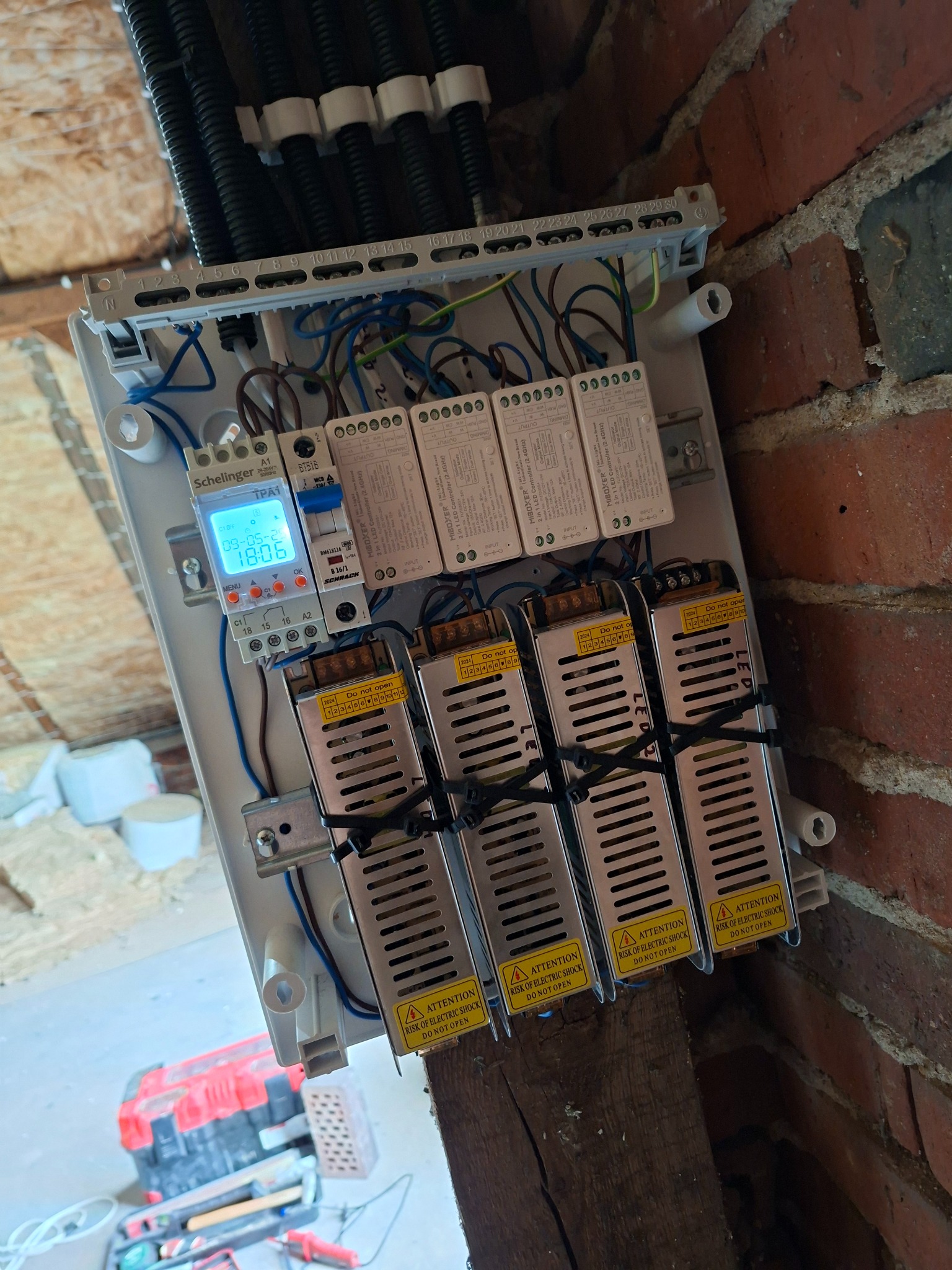 Rozdzielnica elektryczna z przewodami, zasilaczami LED i sterownikiem czasowym zamontowana na ceglanej ścianie i drewnianej belce, widok z dołu.