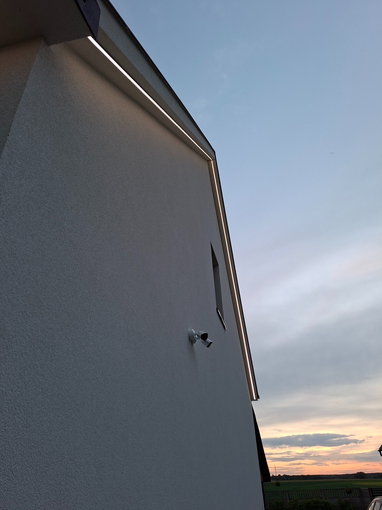 Elewacja domu z oświetleniem LED i kamerą monitoringu na tle wieczornego nieba. Minimalistyczny design, nowoczesne rozwiązania.