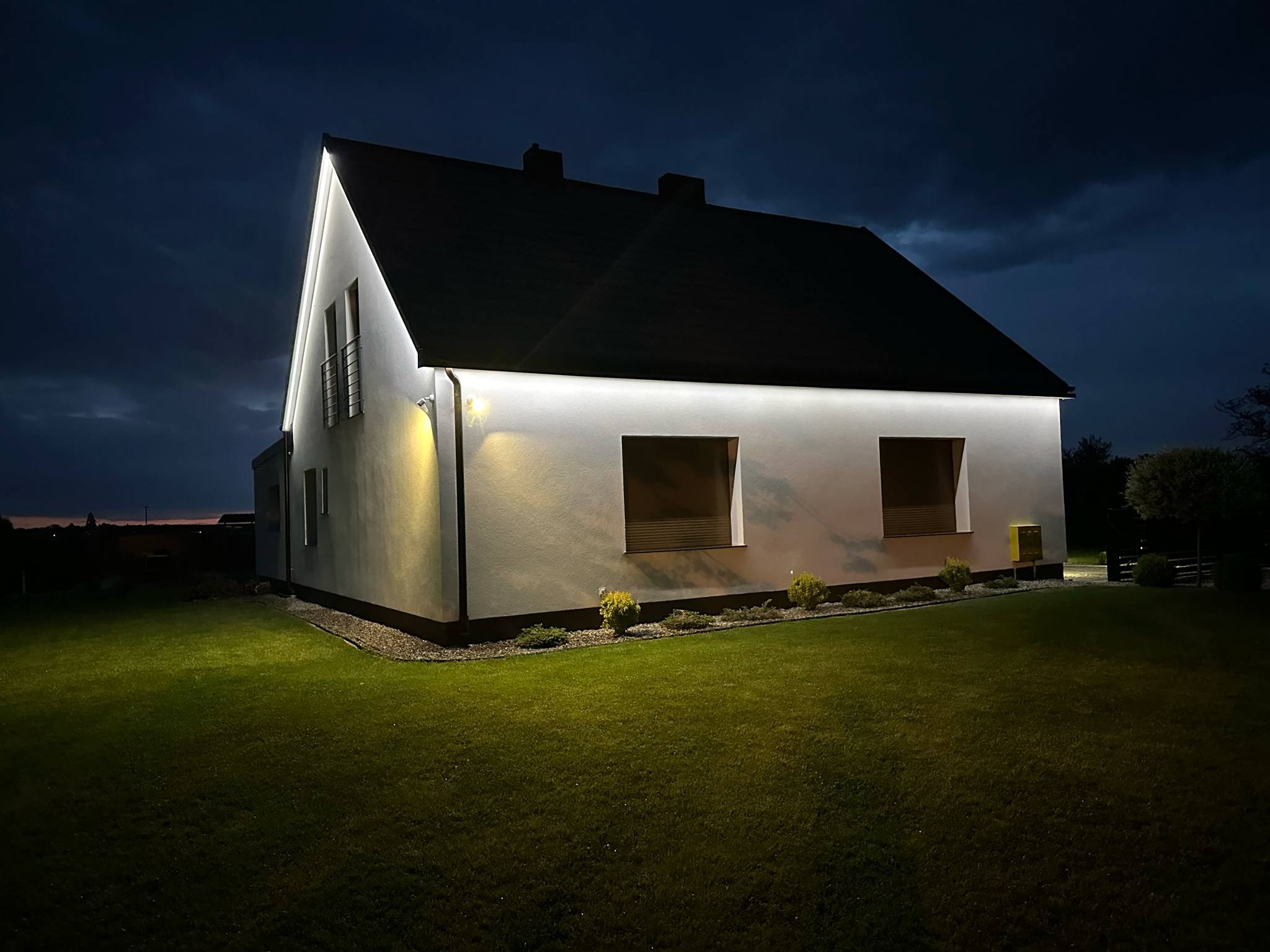Nowoczesny dom jednorodzinny oświetlony nocą białym światłem LED, podświetlony dach i elewacja. Widok z ogrodu, ciemne niebo w tle, zielony trawnik.