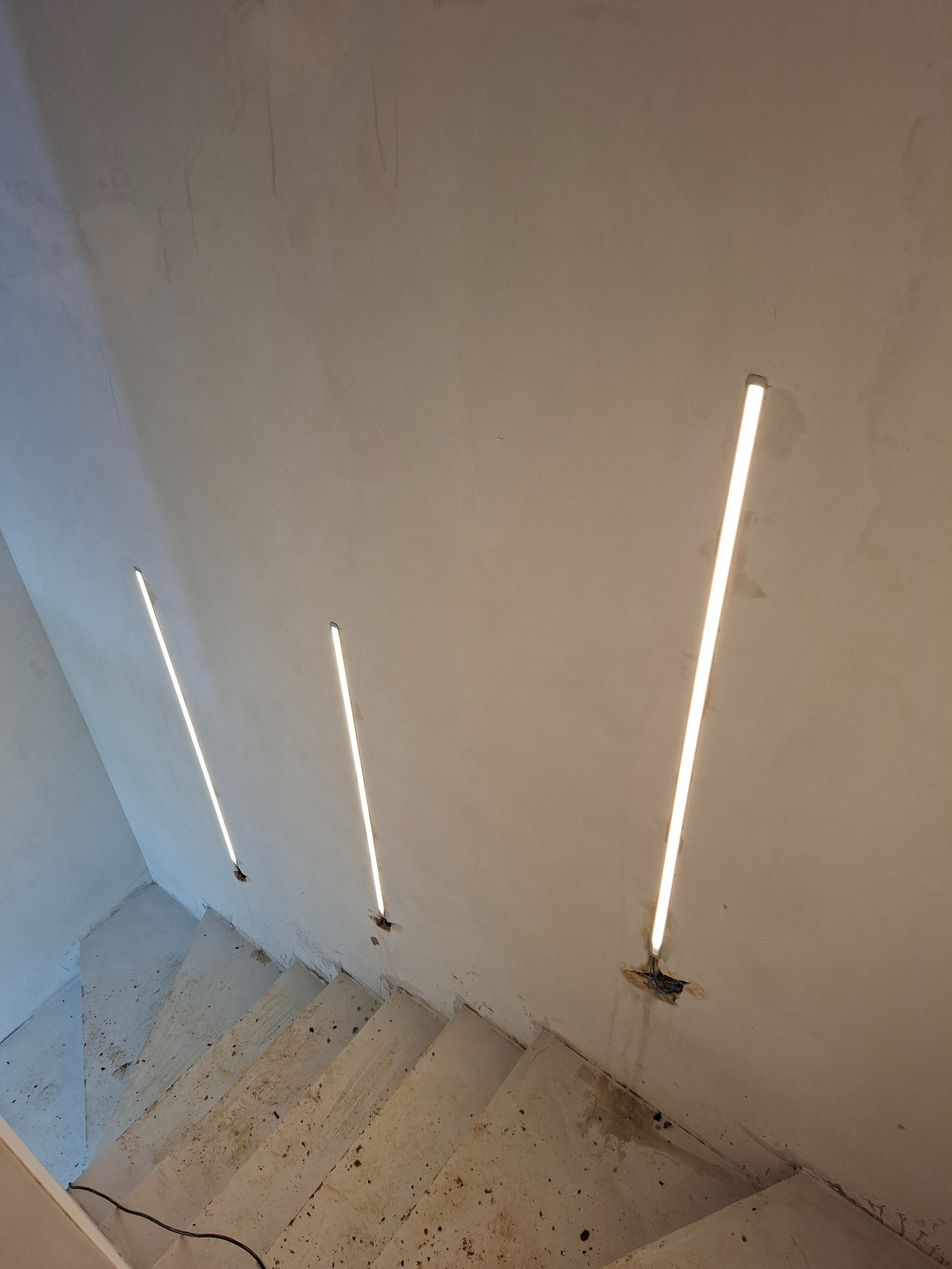 Liniowe oświetlenie LED schodów w Dolsku. Trzy lampy na ścianie, surowy beton, minimalistyczny design. Widok z góry, kabel zasilający widoczny.