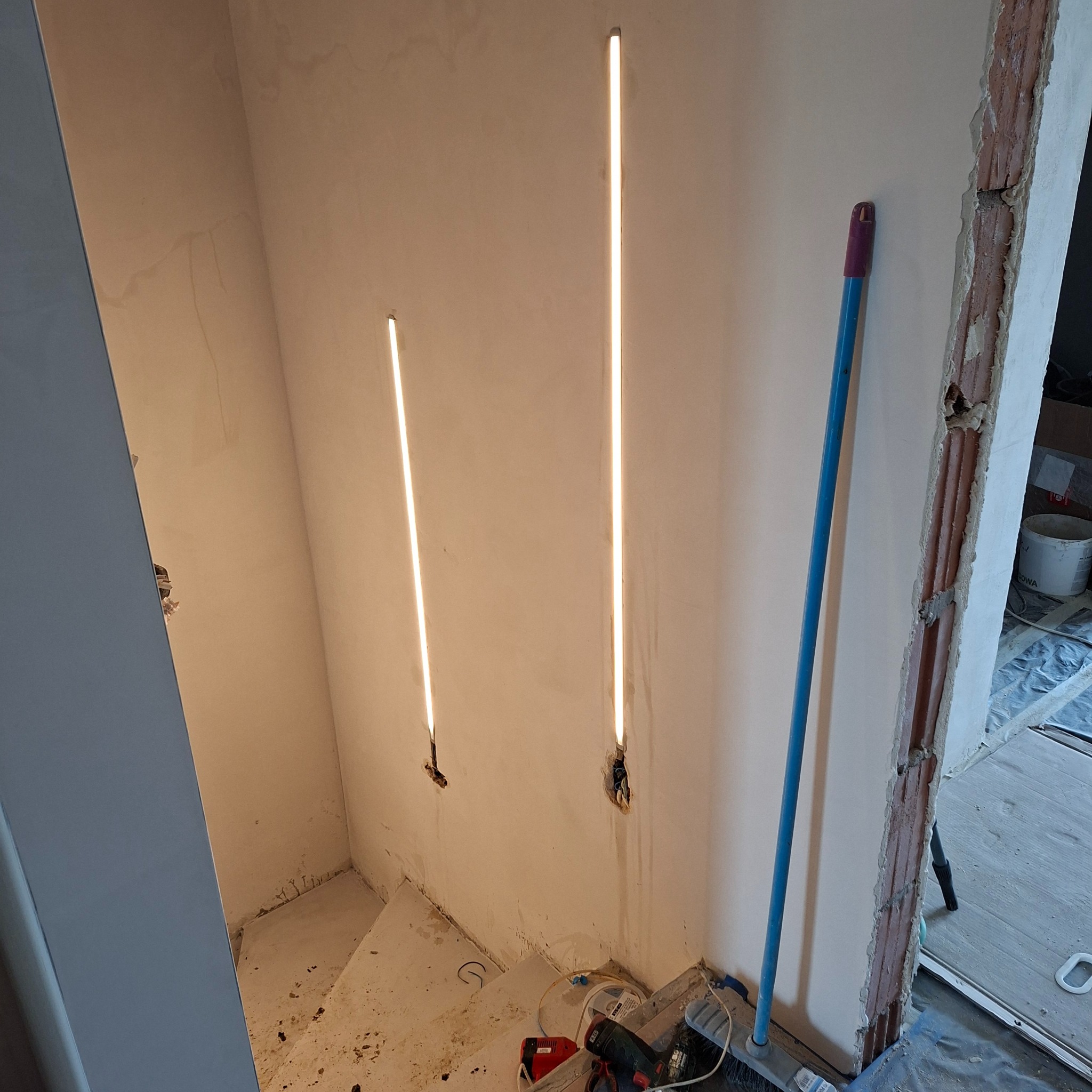 Dwa pionowe, smukłe panele LED w ścianie klatki schodowej w trakcie montażu. Widoczne przewody i narzędzia. W tle surowe wykończenie.