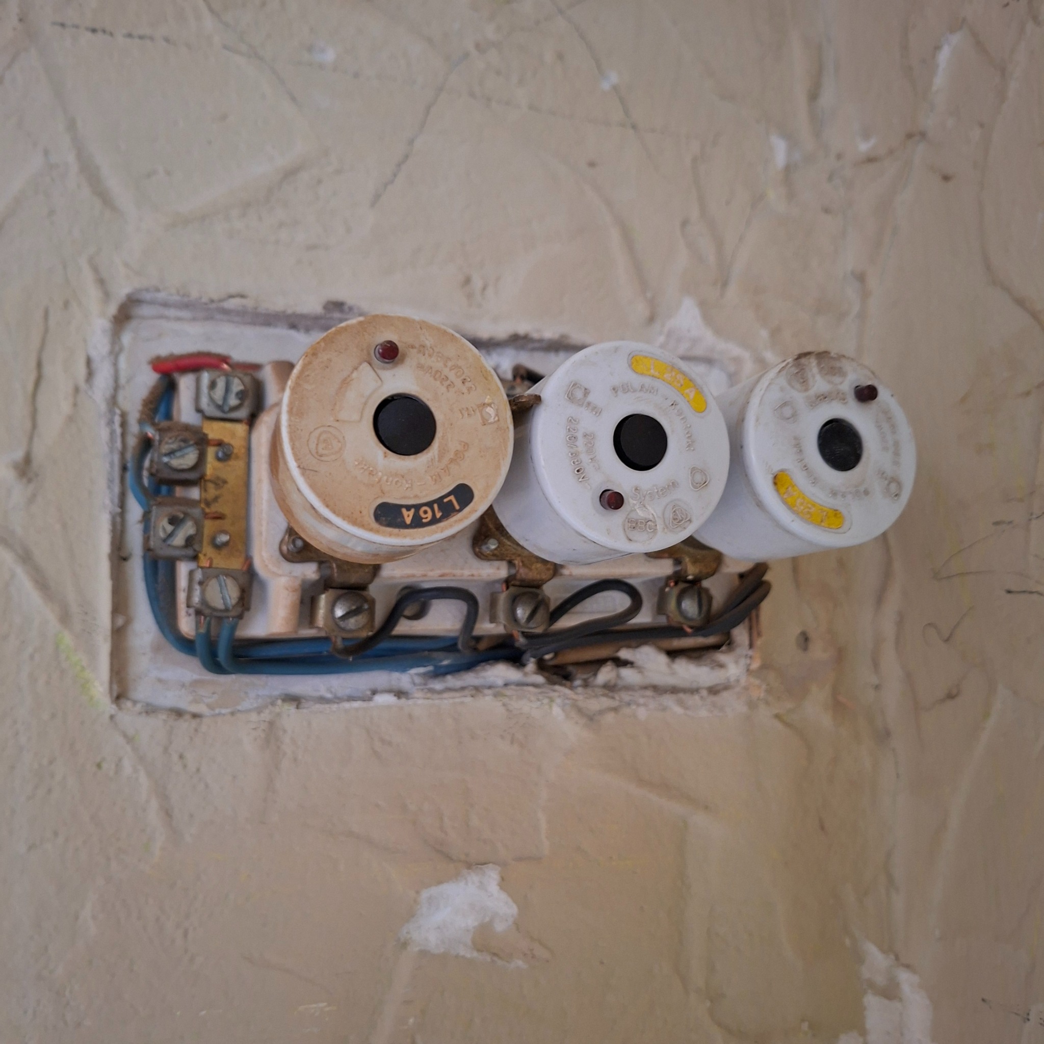 Gniazdo bezpiecznikowe z trzema bezpiecznikami topikowymi na ścianie o nierównej fakturze. Widoczne przewody elektryczne i elementy mocujące.