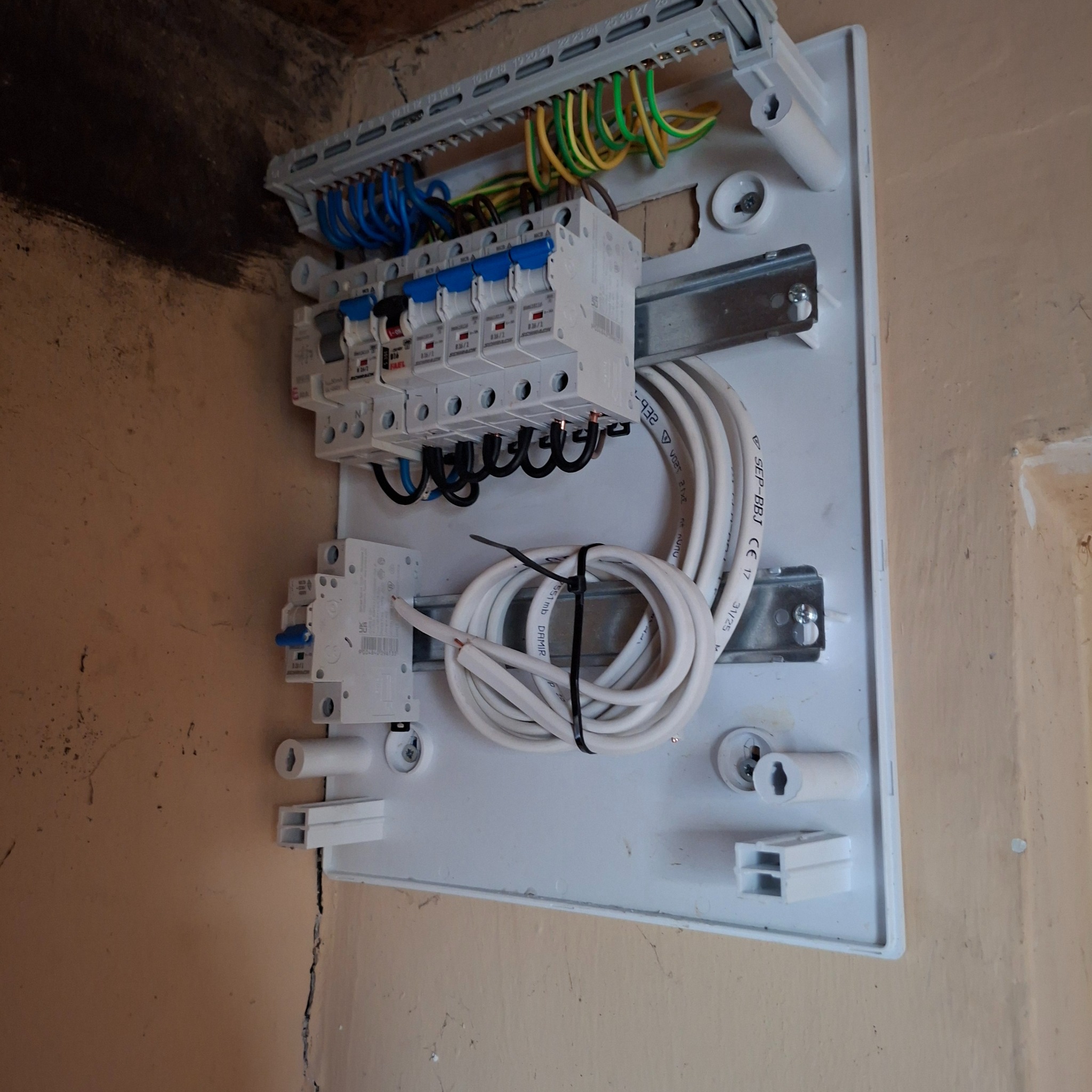 Rozdzielnica elektryczna z bezpiecznikami, przewodami i listwą zaciskową zamontowana na ścianie. Widoczne kable w kolorach: niebieskim, zielono-żółtym i brązowym.