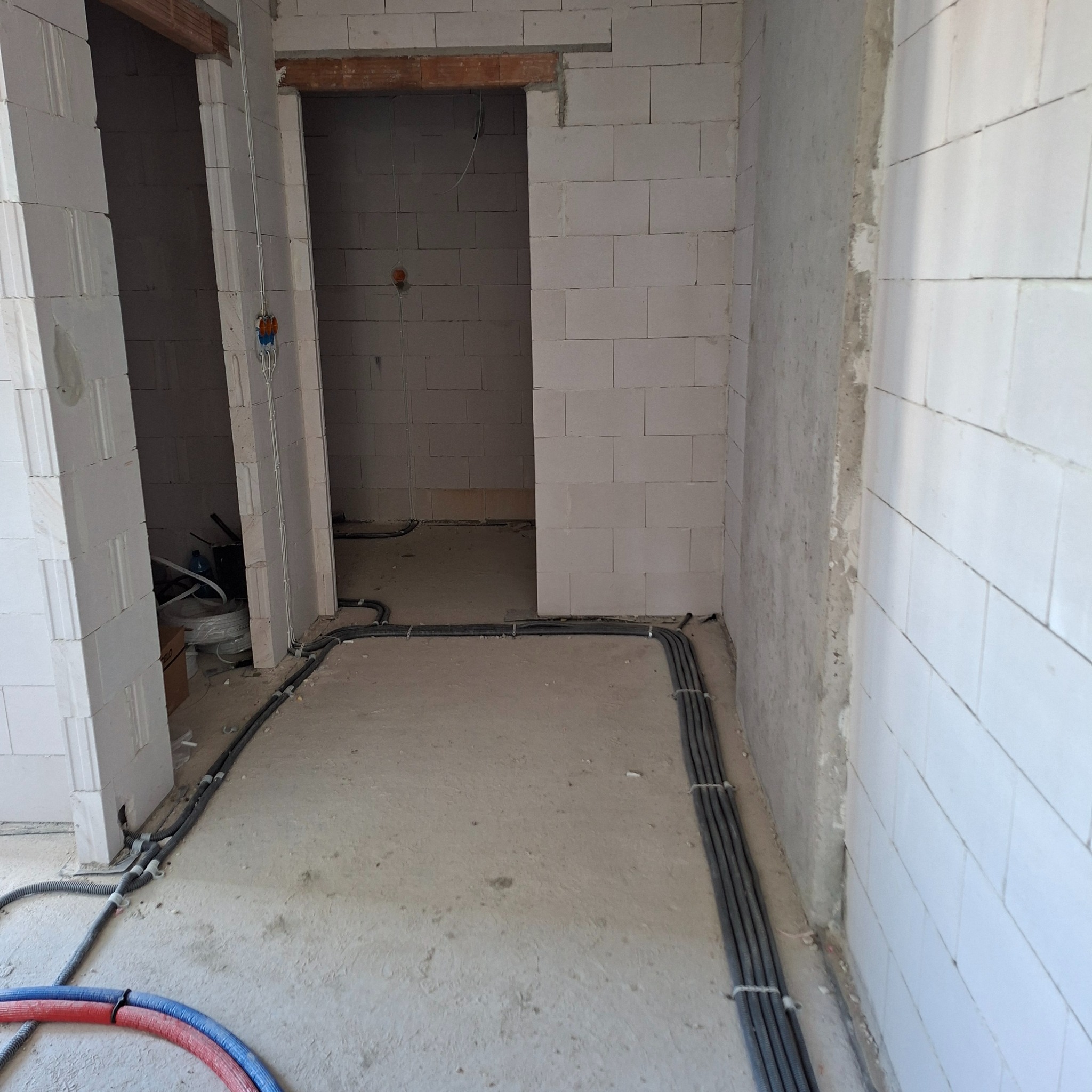 Kable elektryczne w szarych peszlach ułożone wzdłuż ścian w surowym wnętrzu z białego bloczka. W tle widoczne otwory drzwiowe.