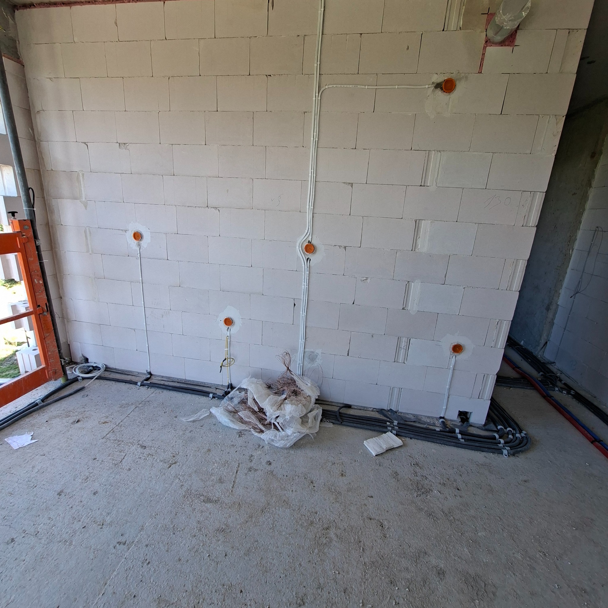 Wnętrze w trakcie prac elektrycznych: białe ściany z bloczków, widoczne peszle z przewodami w ścianach i na podłodze, pomarańczowe puszki instalacyjne.