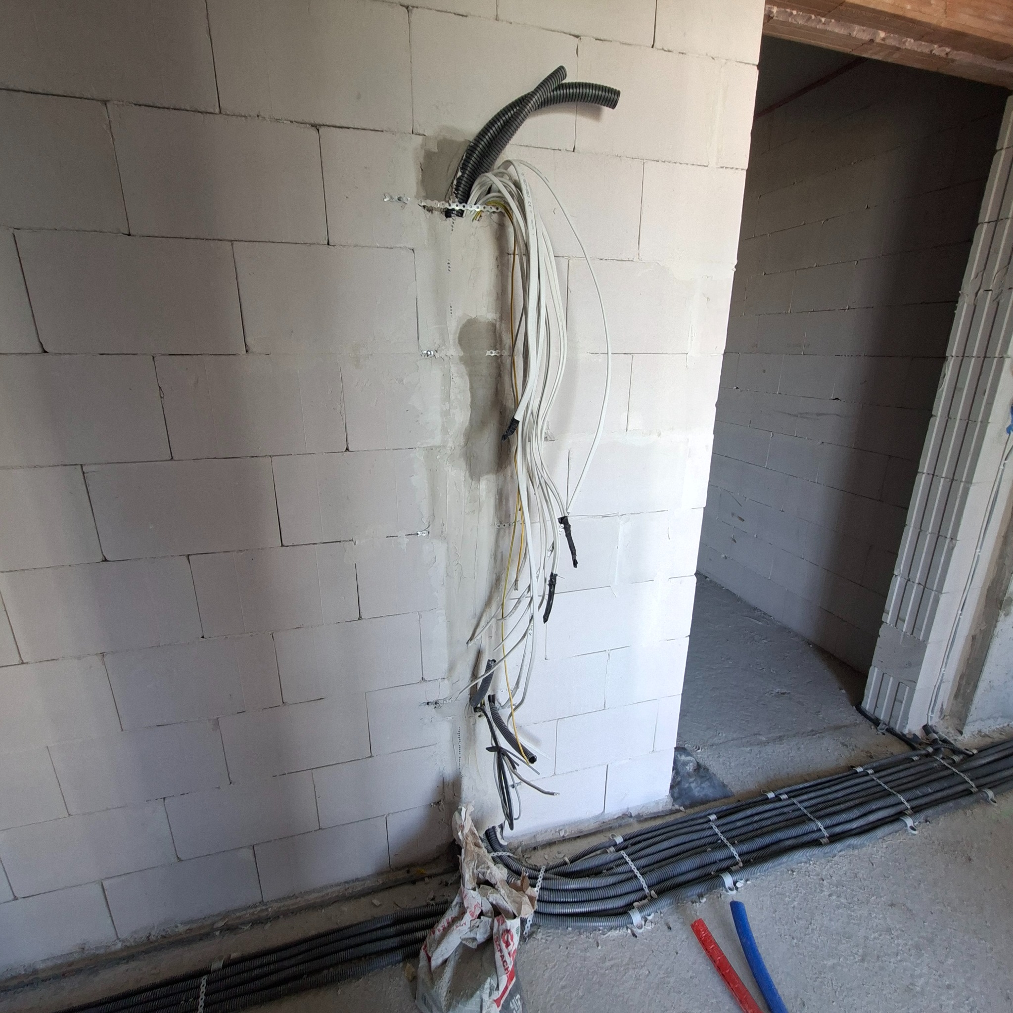 Instalacja przewodów elektrycznych w ścianie z betonu komórkowego. Widoczne kable w peszlach i przewody wychodzące ze ściany, zabezpieczone taśmą. Stan surowy budynku.