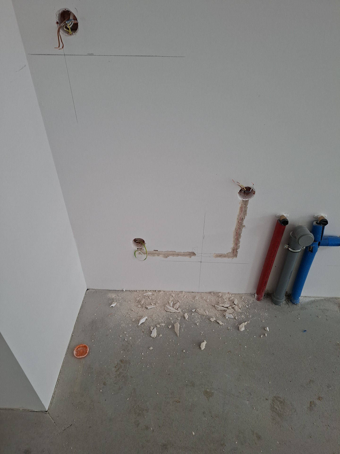 Ściana z widocznymi otworami na instalacje elektryczne i rurami wodnymi w kolorach: czerwonym, szarym i niebieskim. Widoczne ślady po wierceniu.