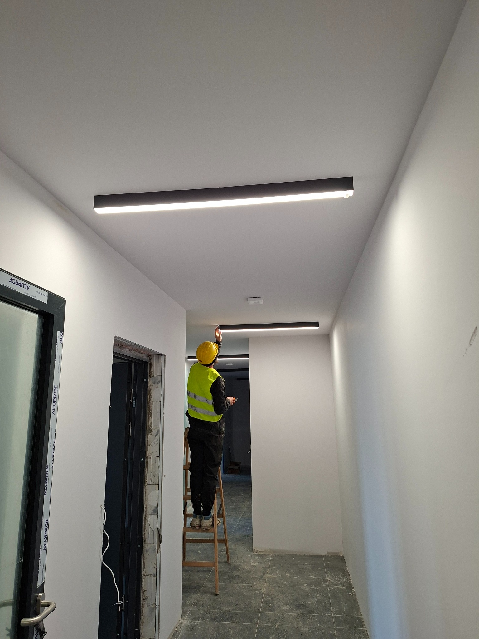 Elektryk na drabinie montuje podłużne lampy LED w korytarzu z białymi ścianami. Widoczne okna i drzwi w stanie surowym. Żółty kask i kamizelka odblaskowa.