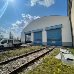 Biały budynek magazynowy z trzema niebieskimi bramami rolowanymi, widoczny z zewnątrz, z torami kolejowymi na pierwszym planie i białym vanem z otwartymi drzwiami po lewej stronie, w słoneczny dzień.