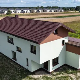 Dwukondygnacyjny dom z brązowym dachem, świeżo otynkowaną elewacją w odcieniach bieli i brązu, widok z góry na tle pól i innych domów.