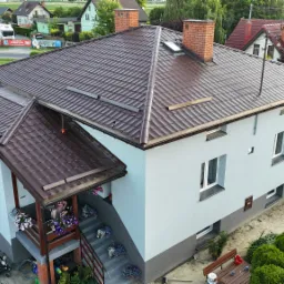 Widok z góry na dom jednorodzinny z nową, brązową dachówką, świeżo pomalowaną elewacją w odcieniach szarości i białym tynkiem, oraz schodami wejściowymi ozdobionymi kwiatami.