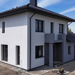 GOLBUD DAWID GOLCZUK SPÓŁKA KOMANDYTOWA - Nowoczesny dom jednorodzinny z biało-szarą elewacją, ciemnym dachem i czarnymi oknami. Charakterystyczny, ciemnoszary element architektoniczny przy wejściu. Teren wokół w trakcie zagospodarowania.