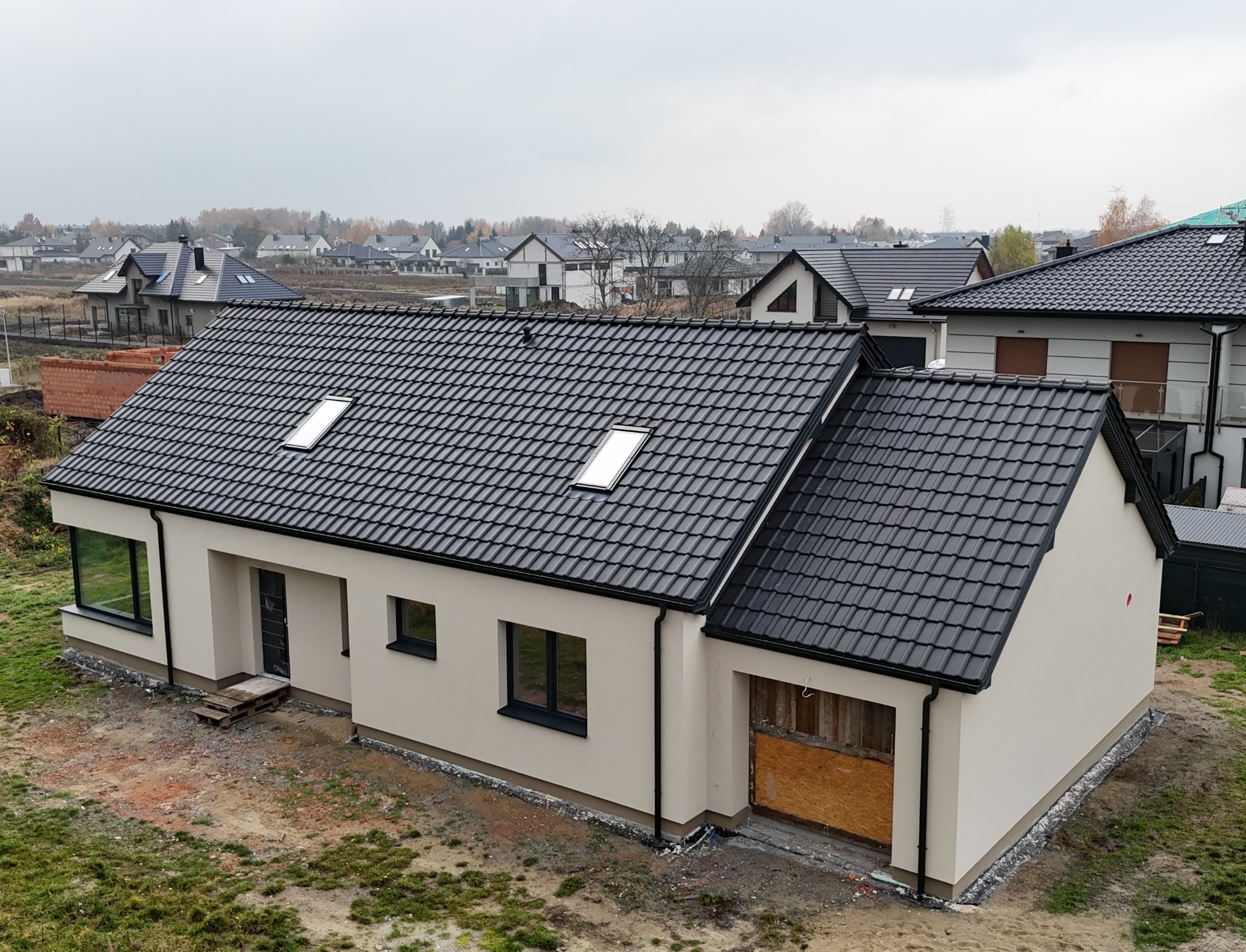 Nowoczesny dom jednorodzinny z grafitową dachówką i jasną elewacją, widok z góry na tle innych domów. Częściowo wykończony teren wokół budynku.