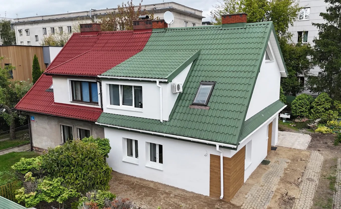 Dom po remoncie elewacji i dachu, z połową dachu w kolorze czerwonym, a drugą w zielonym. Widoczne ocieplenie i nowy tynk na jednej części budynku.