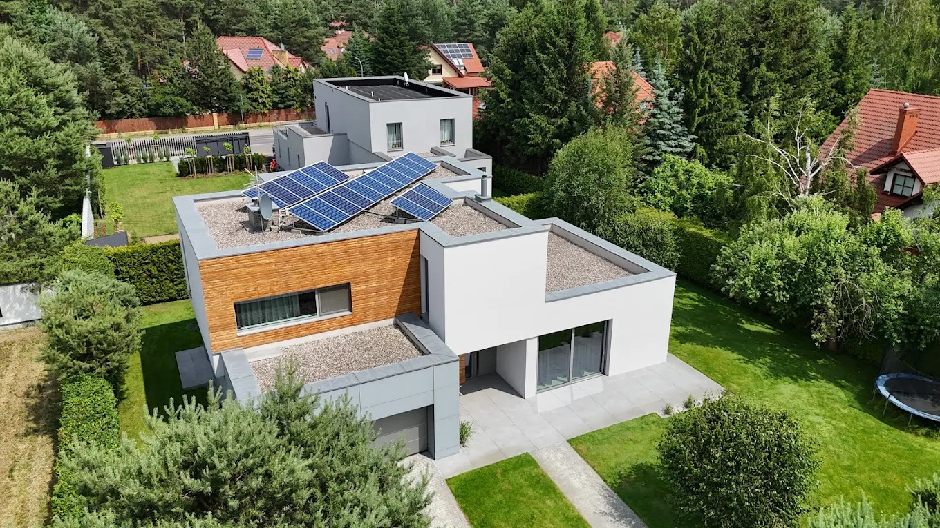 Nowoczesny dom z panelami słonecznymi na płaskim dachu, drewnianą elewacją i białymi ścianami. Widok z góry, otoczony zielenią.