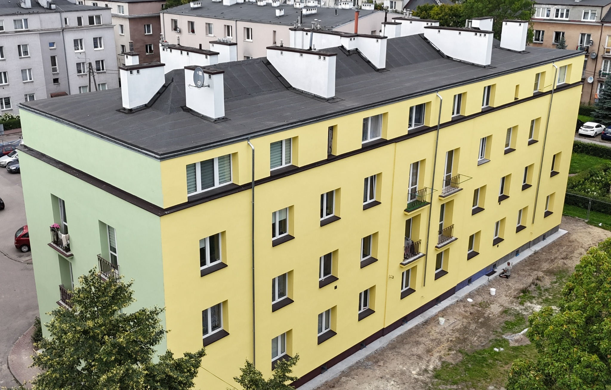 Odnowiona elewacja bloku w odcieniach żółci i zieleni, z balkonami i nowymi oknami. Widok z góry ukazuje detale wykończenia i otoczenie budynku.