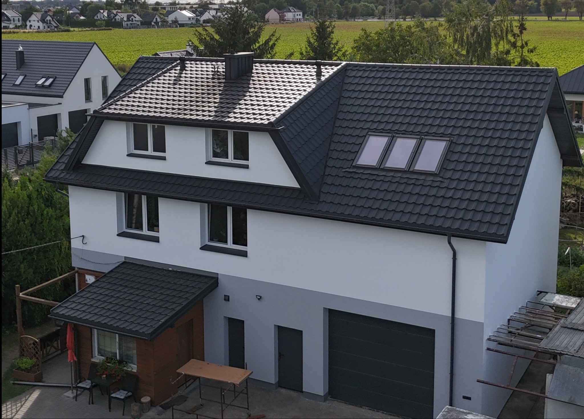 Dwukondygnacyjny dom z ciemnoszarą dachówką i jasną elewacją, widziany z góry. Widoczne okna dachowe, garaż i fragment ogrodu. W tle pola i zabudowa wiejska.