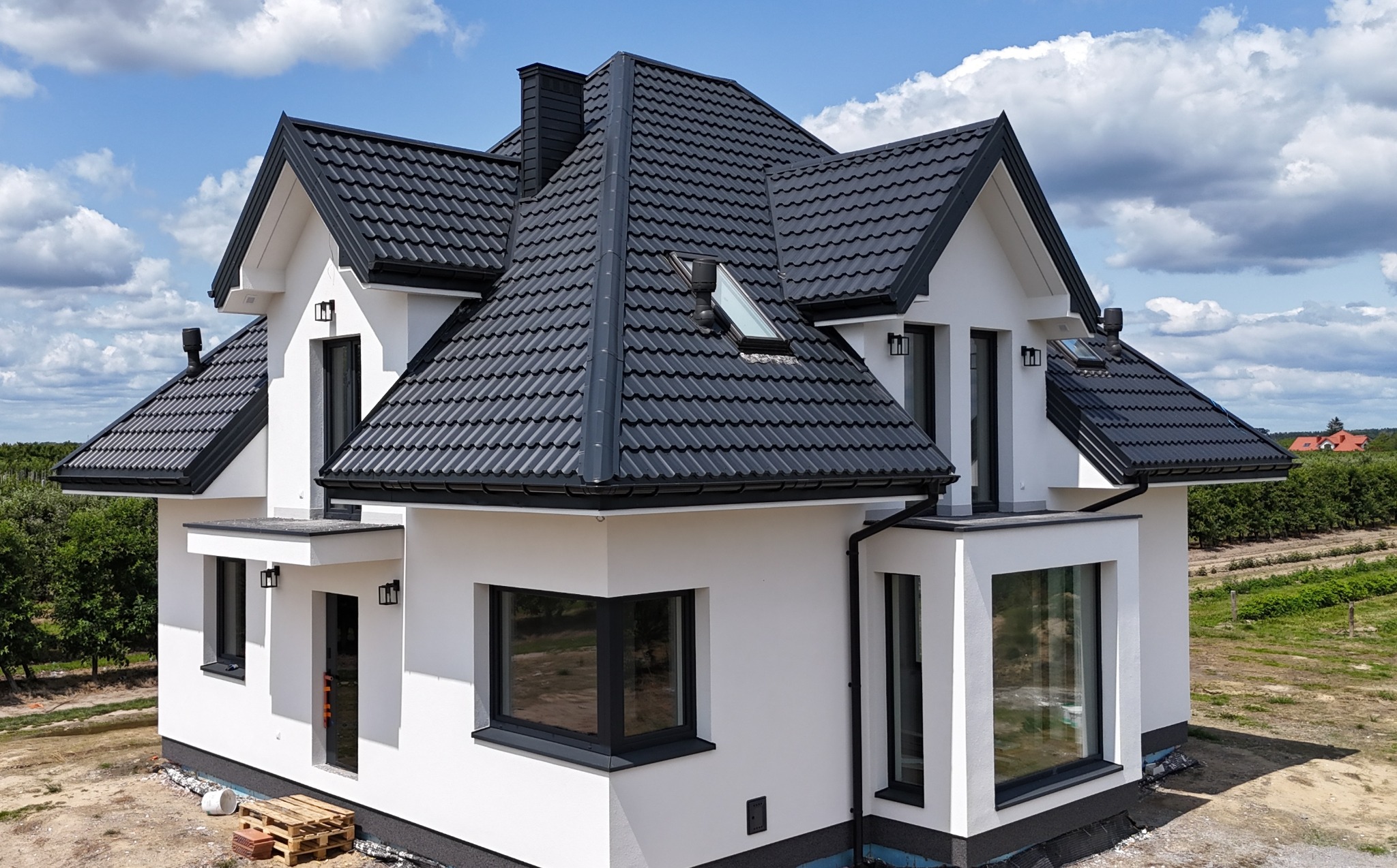 Nowoczesny dom jednorodzinny z czarnym dachem z blachodachówki i białymi ścianami, widok z zewnątrz, w tle błękitne niebo z chmurami.