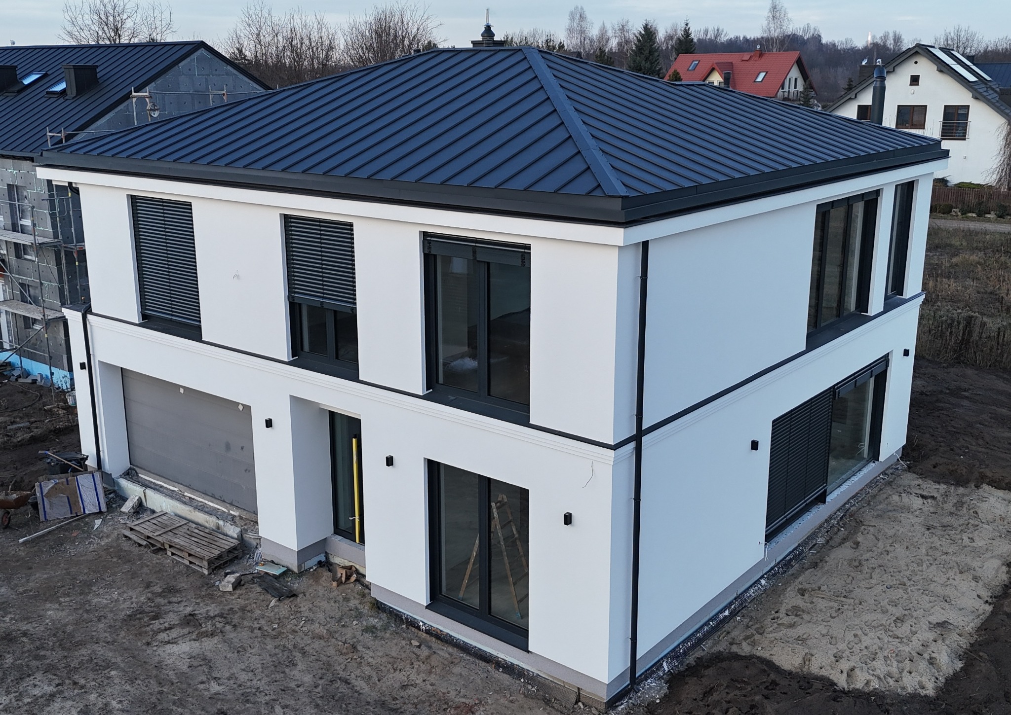 Nowoczesny, dwukondygnacyjny dom z białą elewacją i ciemną, metalową dachówką. Widoczne czarne ramy okienne i żaluzje. Teren wokół domu częściowo zagospodarowany.