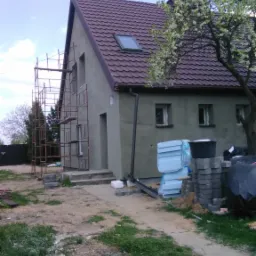 Dom jednorodzinny w trakcie prac wykończeniowych elewacji, widoczne rusztowania, ocieplenie styropianem i przygotowane materiały budowlane na posesji, w tle kwitnące drzewo.