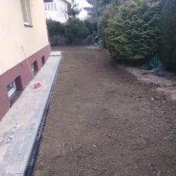 Przygotowanie terenu pod ocieplenie ścian w Piastowie: widok na odsłonięty fundament, ułożoną kostkę brukową z odpływem liniowym i leżącą miotłę przy ścianie budynku.