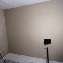 Świeżo pomalowana ściana w pokoju, zabezpieczona podłoga, widoczna drabina i lampa halogenowa na statywie.