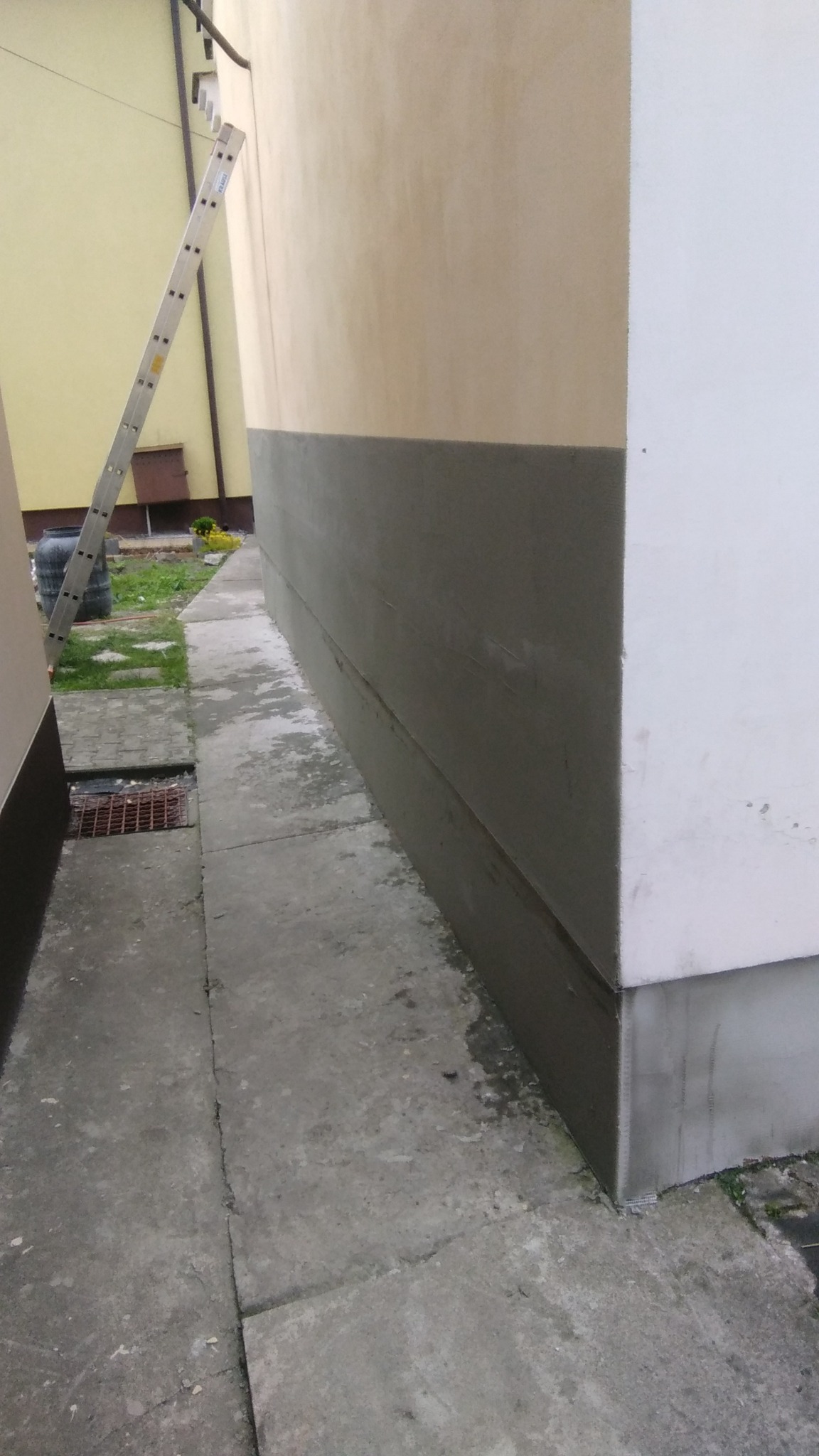 Ściana budynku z częściowo wykonanym ociepleniem, widoczna drabina oparta o ścianę, szara podmurówka i jasna elewacja powyżej, obok chodnik z kostki betonowej.