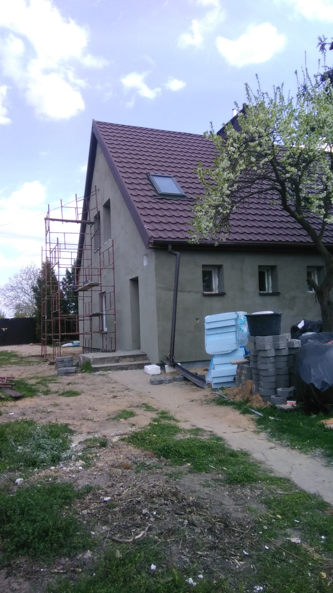 Dom jednorodzinny w trakcie prac wykończeniowych elewacji, widoczne rusztowania, ocieplenie styropianem i przygotowane materiały budowlane na posesji, w tle kwitnące drzewo.